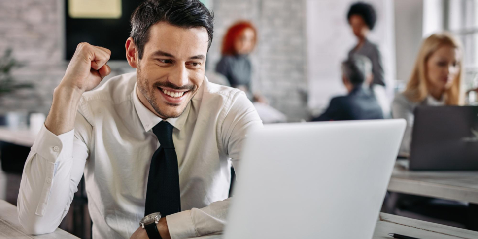 young-happy-businessman-using-computer-office-celebrating-good-news-there-are-people-background